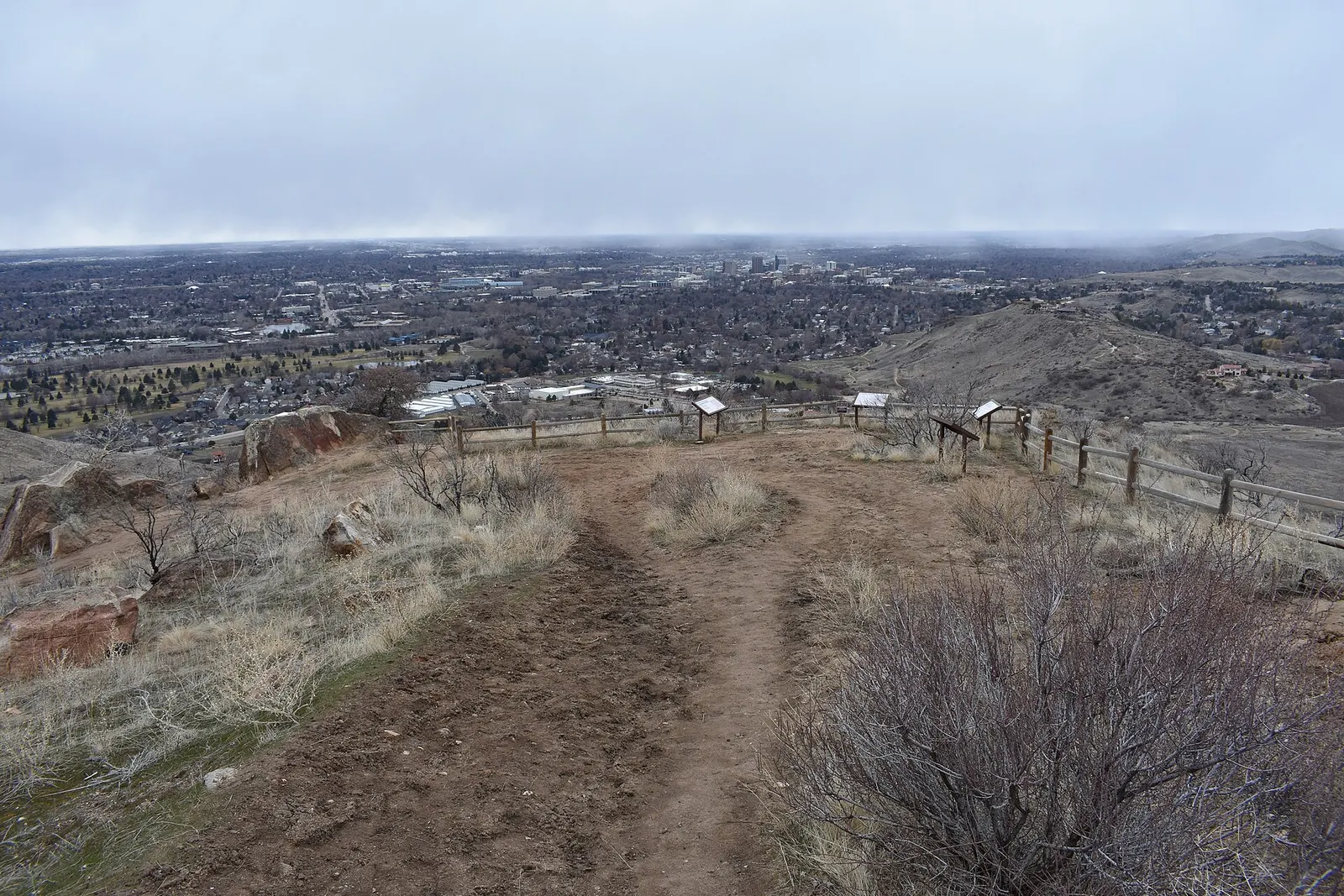 View from a dirt trail overlook on the Boise foothills across the city under low gray clouds, with trailside signs along a wooden fence and downtown Boise in the distance
