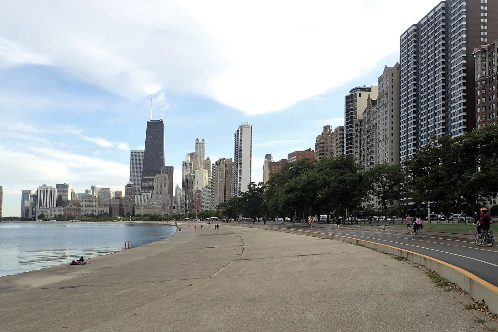 The Chicago skyline rising beyond the Lakefront Trail and Lake Michigan shoreline, with a few cyclists and walkers on the path under a partly cloudy sky