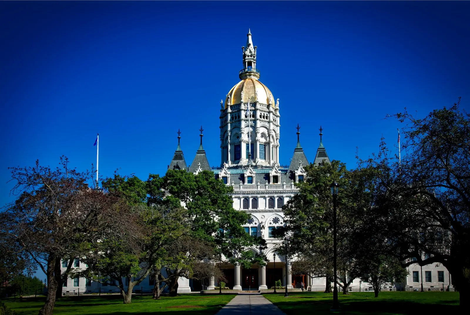 The Connecticut State Capitol in Hartford: a Gothic-style building with a gold dome and spire under a deep blue sky, seen along a path between trees