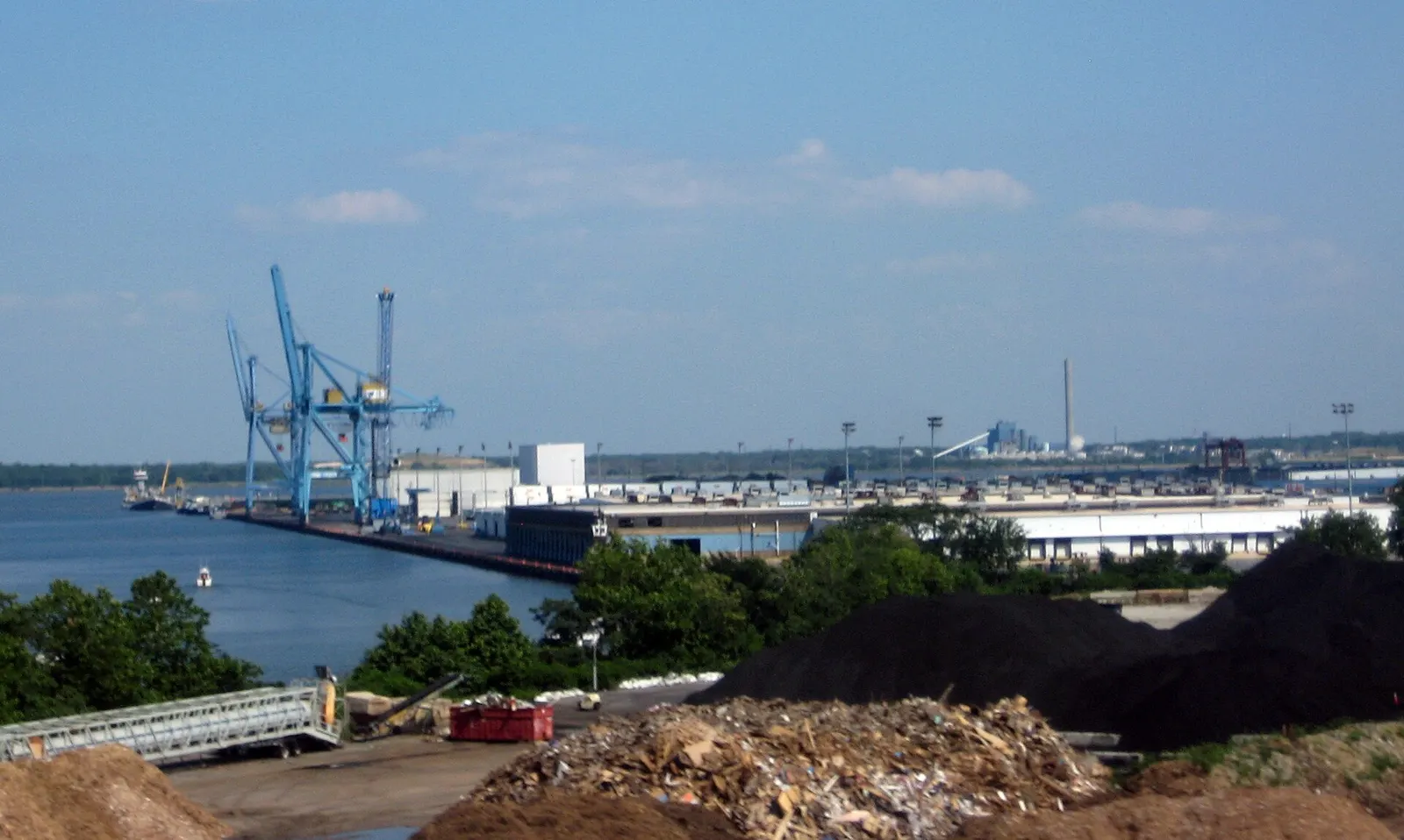 Container cranes and warehouses along the Port of Wilmington waterfront on the Delaware River, with piles of mulch and debris in the foreground under a hazy blue sky