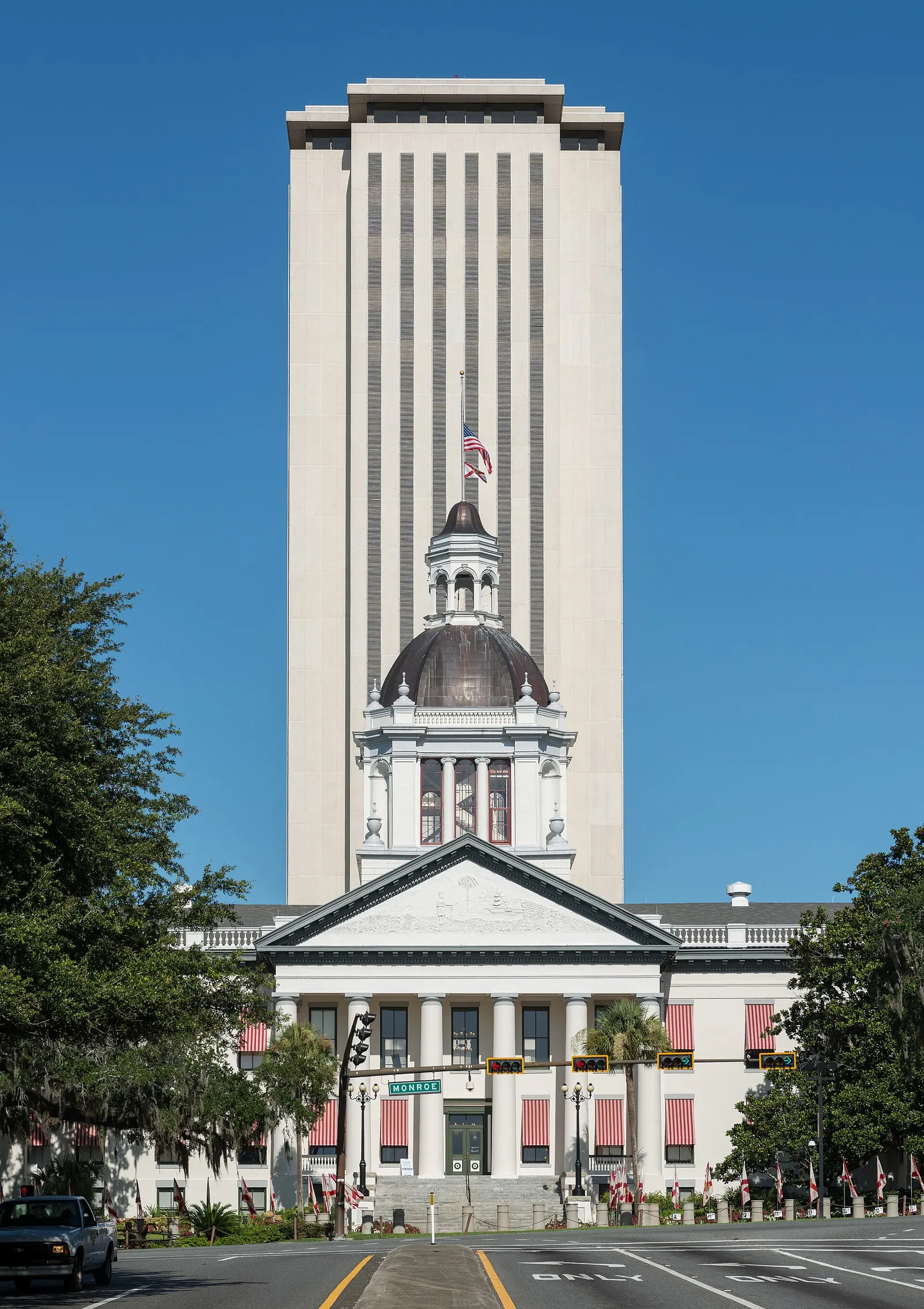 The white columned Old Florida Capitol with a copper dome and red-striped awnings in front of the tall New Capitol tower in Tallahassee, seen from the street under a blue sky