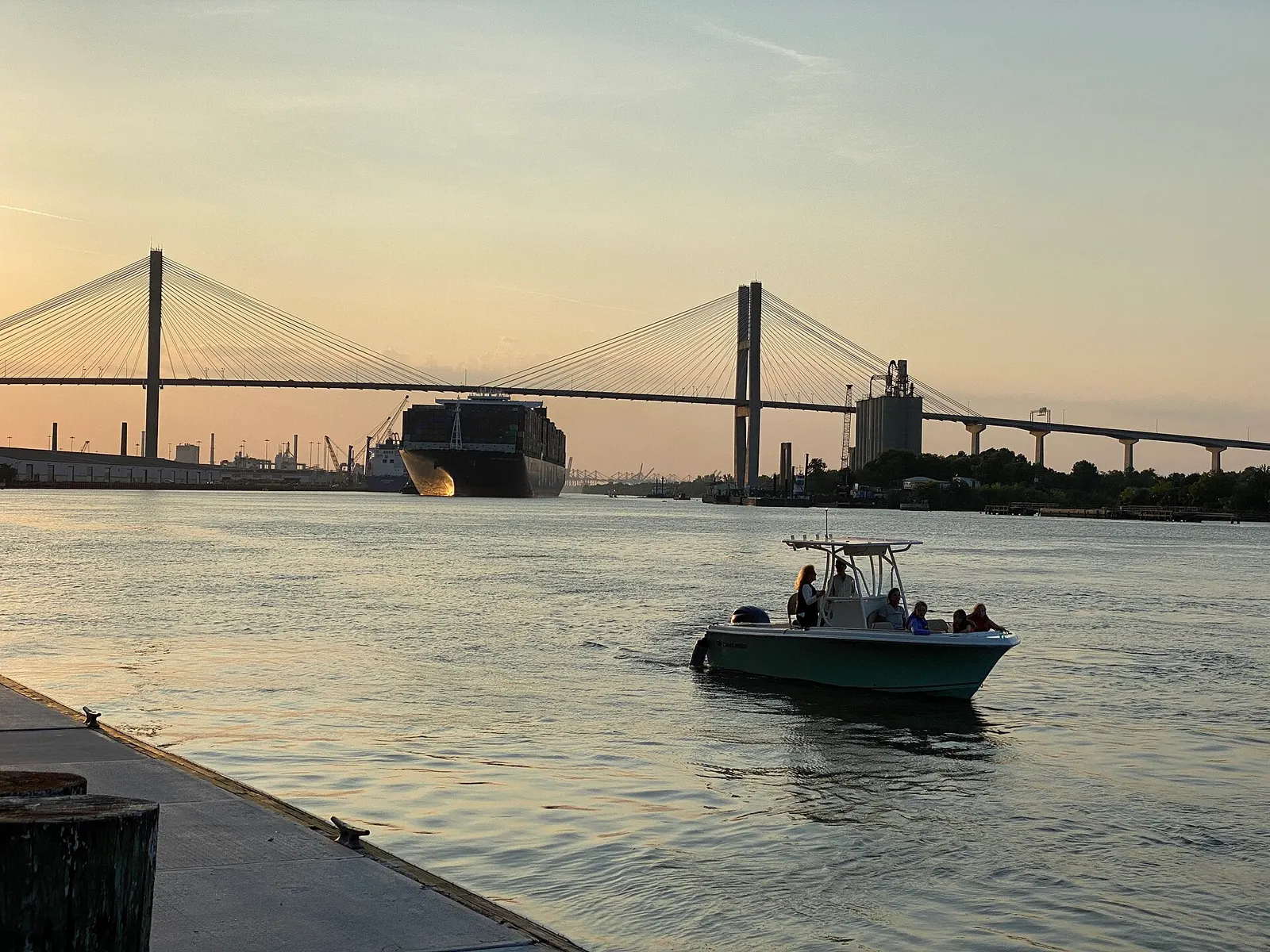 A large container ship approaching the cable-stayed Talmadge Memorial Bridge on the Savannah River at sunset, with a small motorboat in the foreground and a riverfront dock at left