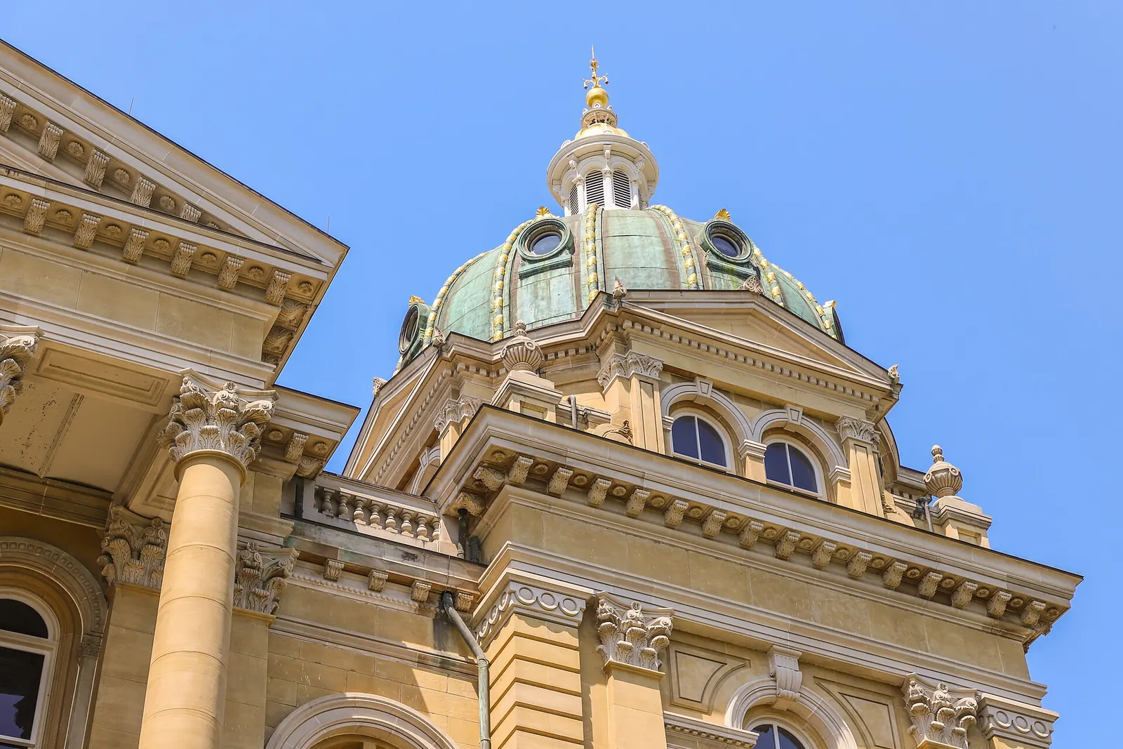 The green and gold dome of the Iowa State Capitol above carved stone columns against a clear blue sky