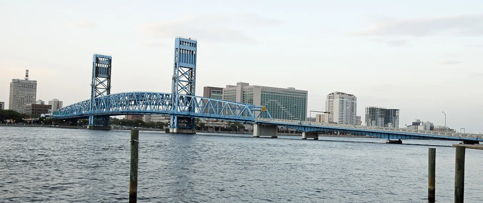 The blue steel Main Street Bridge crossing the St. Johns River in Jacksonville, with downtown buildings on the far bank at dusk.