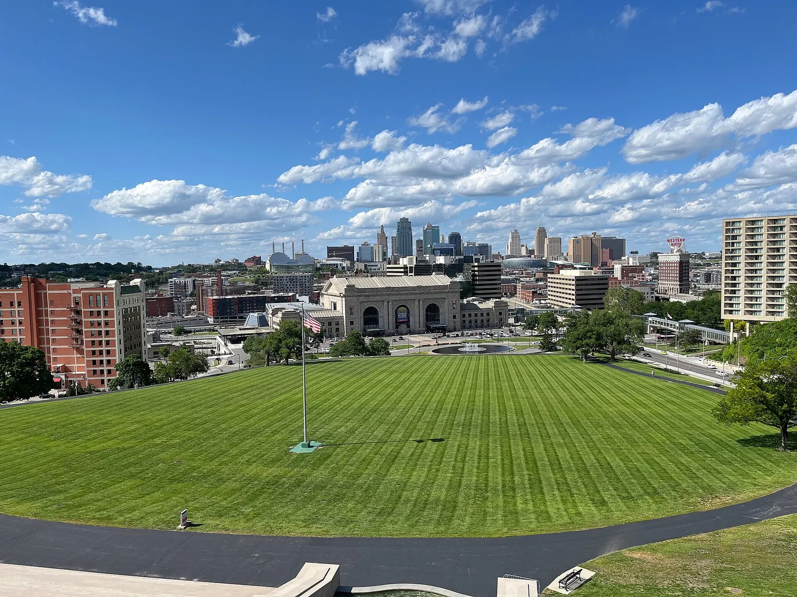 A view across a broad mowed lawn toward a large stone building with arched entrances, with a downtown skyline behind it under a blue sky with clouds.
