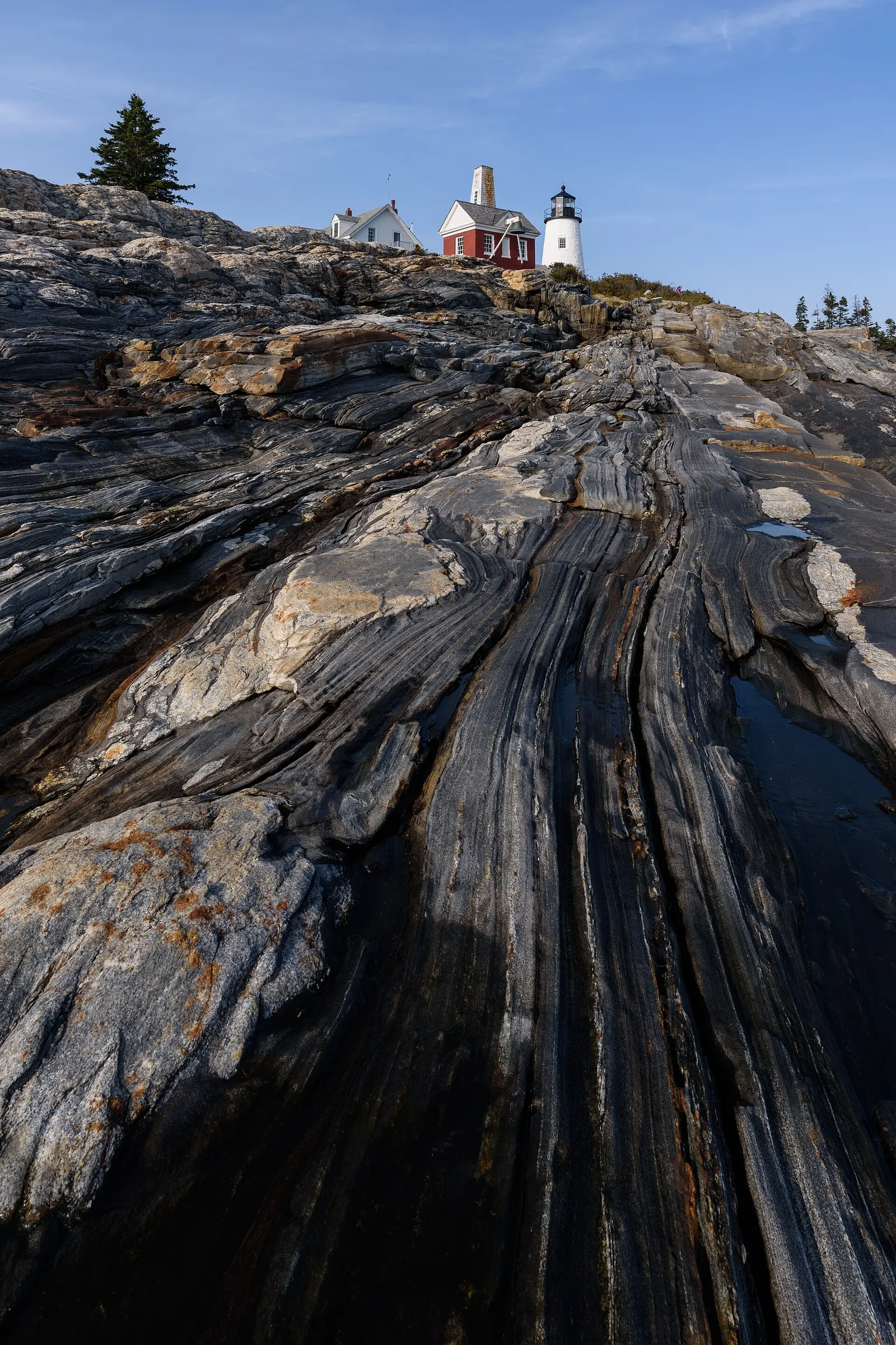 A white lighthouse tower and a red-roofed keeper house on a rocky rise at Pemaquid Point, Maine, seen from below across banded gray and white striped rock