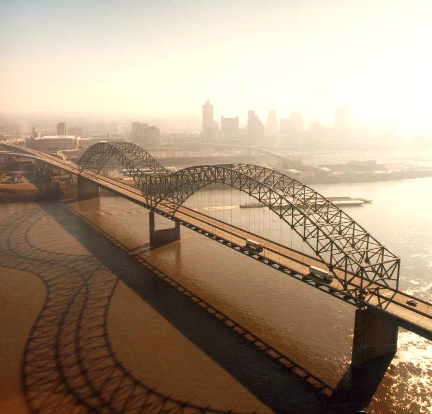 Aerial view of the steel arches of the Hernando de Soto Bridge over the brown Mississippi River at Memphis, with trucks on the deck, a barge below and the downtown skyline in haze