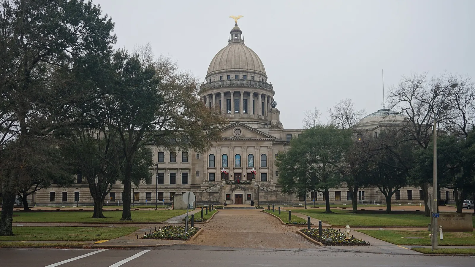 A stone capitol building with a large dome and a pedimented center section, seen across a lawn on an overcast day with trees on both sides