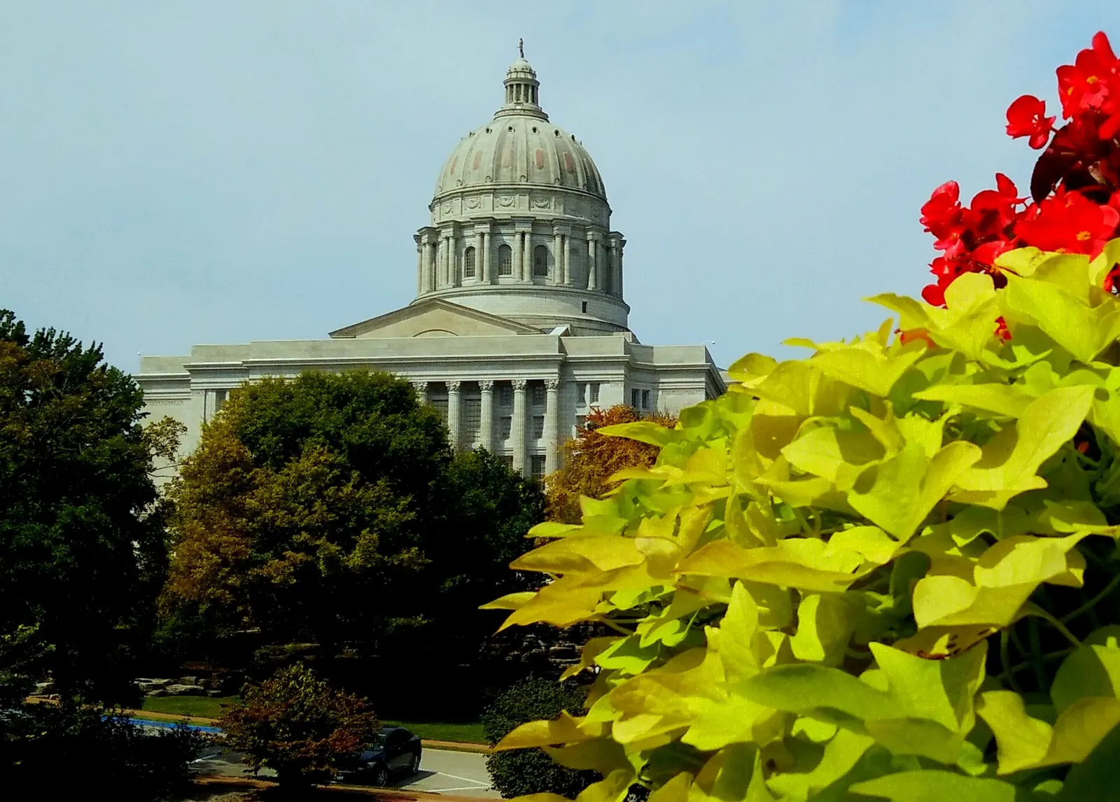 The domed Missouri State Capitol in Jefferson City seen over trees, with red flowers and lime-green foliage in the foreground