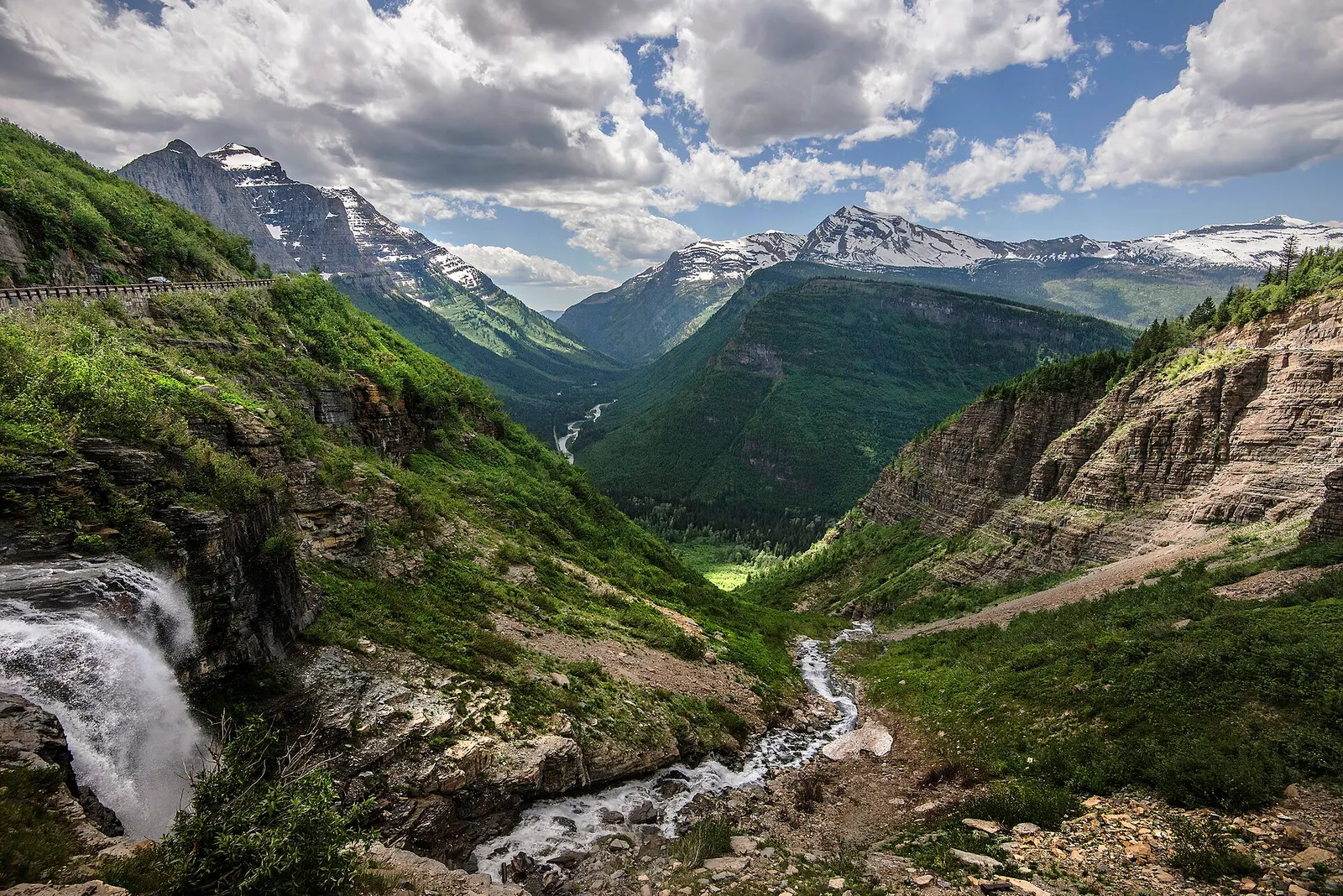 A waterfall and stream drop into a green glacial valley in Glacier National Park, with snow-dusted peaks above and a mountain road cut into the cliff at upper left
