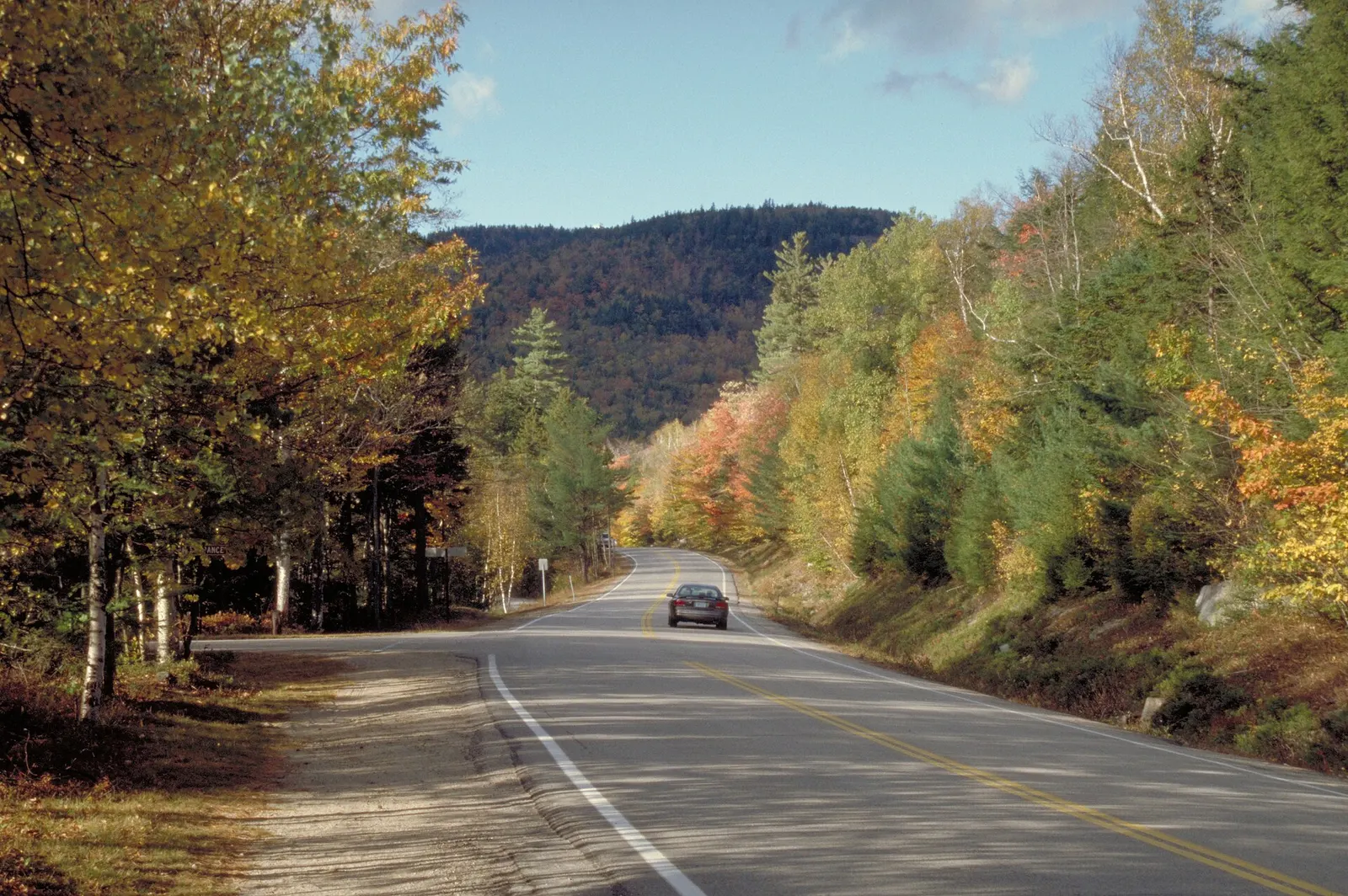A two-lane road on the Kancamagus Highway curving through hardwood forest in fall color, with yellow and orange leaves, a forested hill ahead and a single car on the road