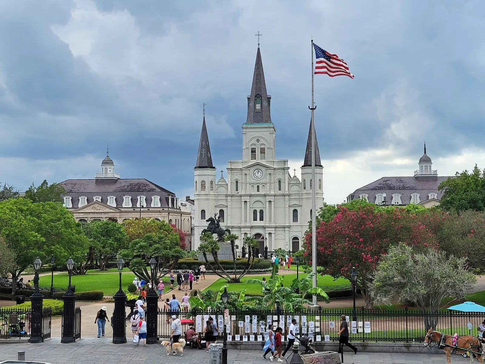 A white three-spired church seen across a small park with a statue, palm fronds and flowering trees, with a flag on a tall pole and dark clouds behind.
