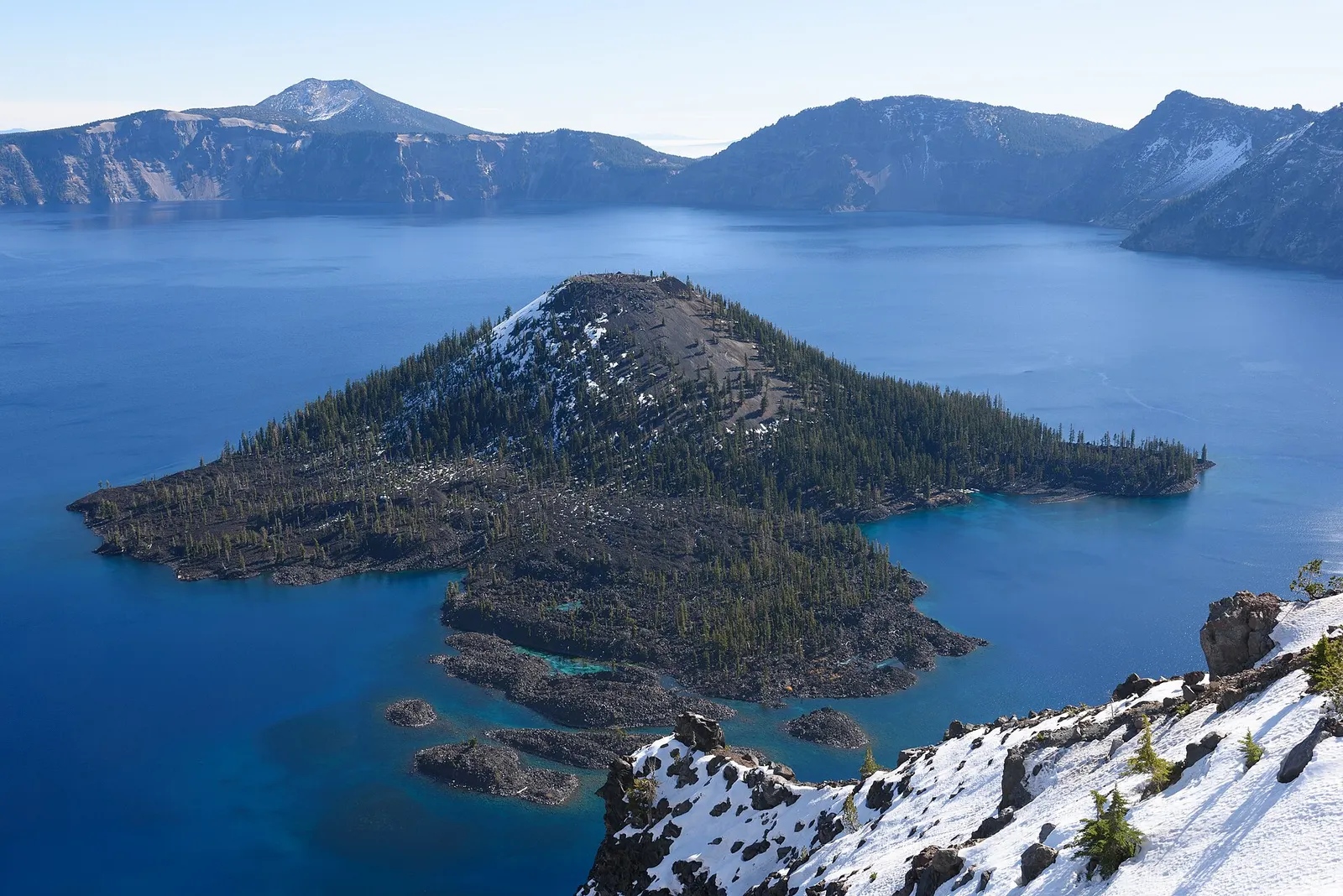 Wizard Island, a forested volcanic cone in the deep blue water of Crater Lake, with snow on the rim rocks in the foreground and caldera walls behind