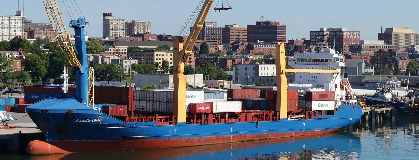 A blue cargo ship named Skogafoss loaded with containers and fitted with two yellow cranes, tied up at a Portland, Maine pier with the downtown brick buildings behind it