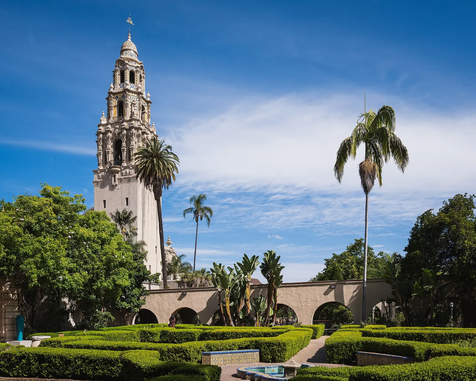 The ornate California Tower in Balboa Park, San Diego, rising above palm trees, clipped hedges and an arcade under a blue sky