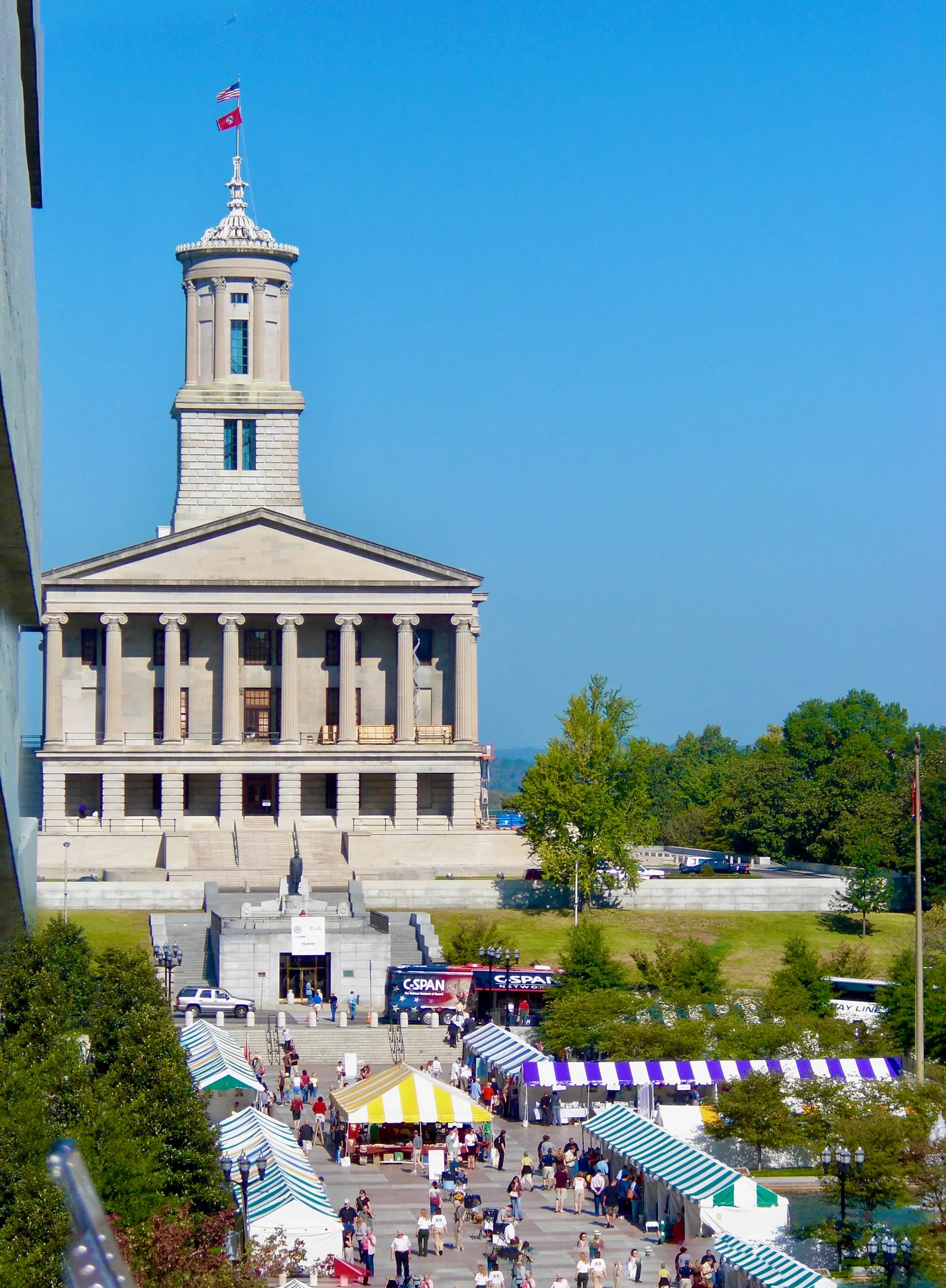 The Tennessee State Capitol in Nashville, a columned stone building with a square tower, seen from above with market tents on the plaza below and a blue sky behind