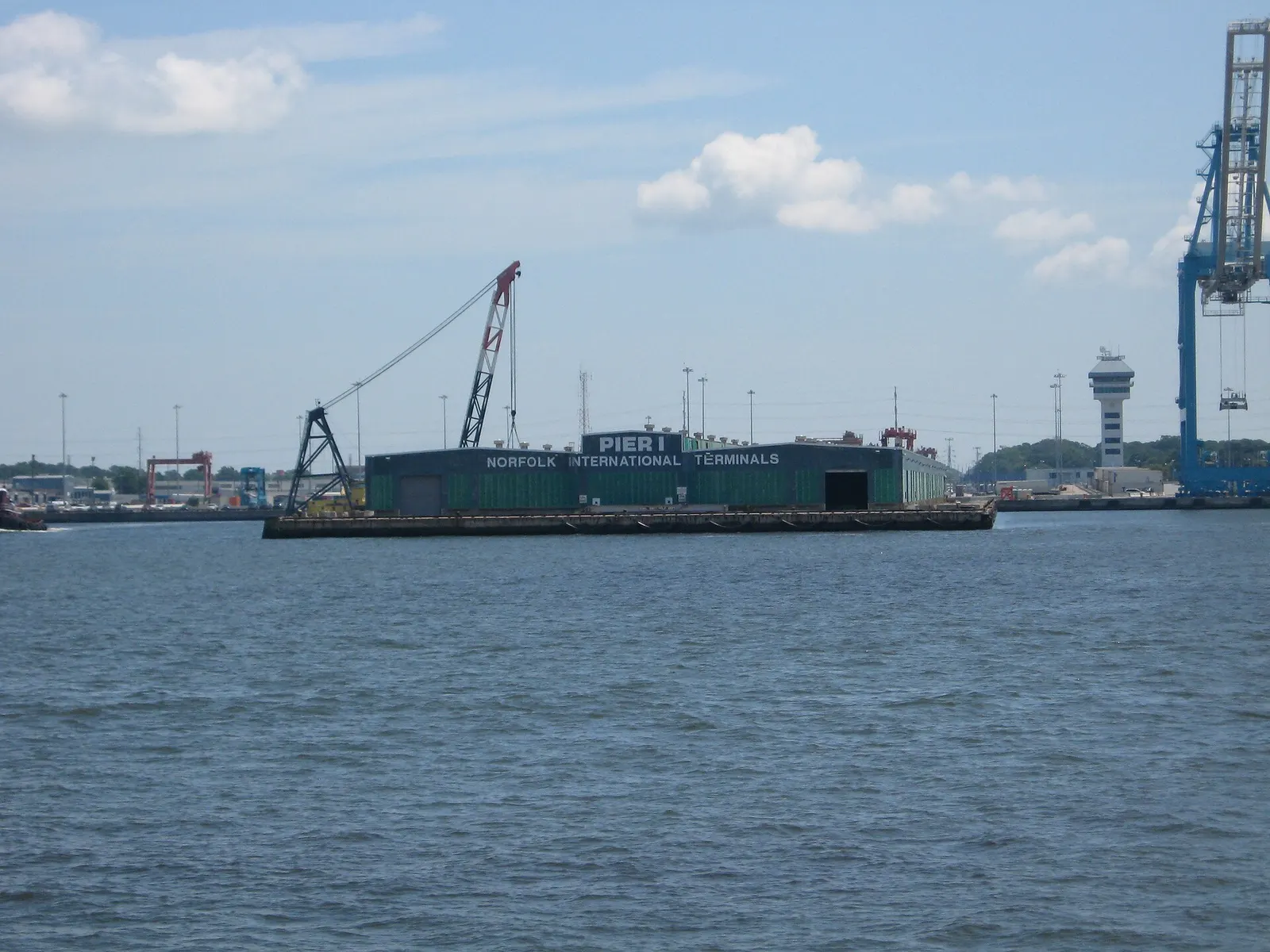 A green cargo shed marked Norfolk International Terminals on a pier seen across the water, with a red crane to its left and a blue container crane at the edge of the frame